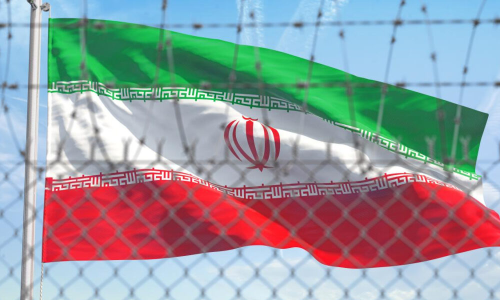 Iranian flag with green, white, and red bands waving behind a chain-link fence with barbed wire against a blue sky (emblem centered).