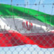 Iranian flag with green, white, and red bands waving behind a chain-link fence with barbed wire against a blue sky (emblem centered).
