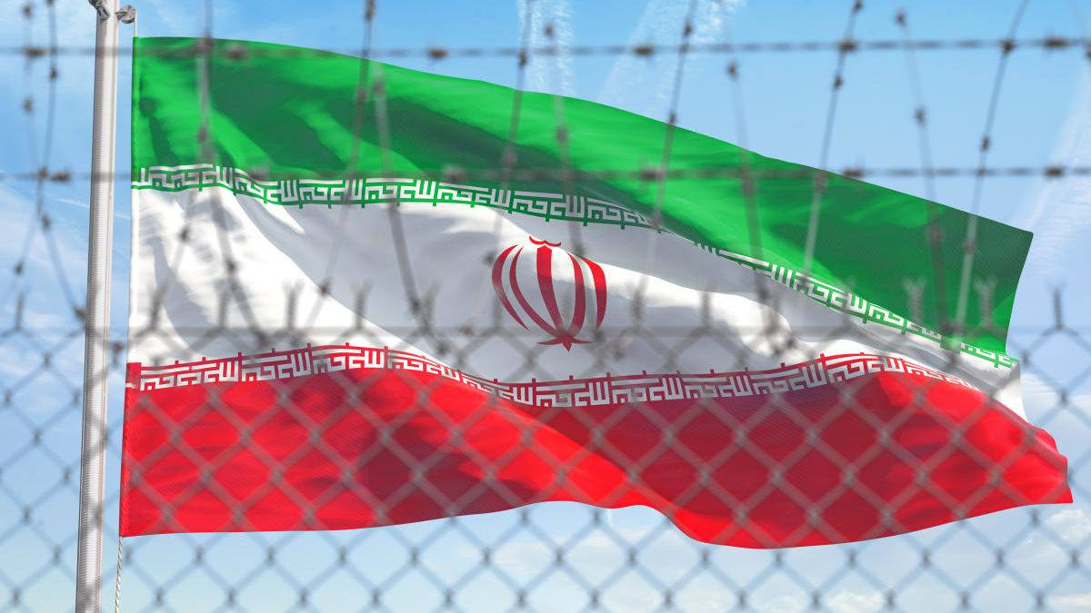 Iranian flag with green, white, and red bands waving behind a chain-link fence with barbed wire against a blue sky (emblem centered).