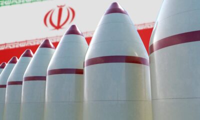 Row of white missiles with red tips and bands, lined up with Iran's flag and emblem in the background, blurred behind them, conveying a weapons display.