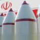 Row of white missiles with red tips and bands, lined up with Iran's flag and emblem in the background, blurred behind them, conveying a weapons display.