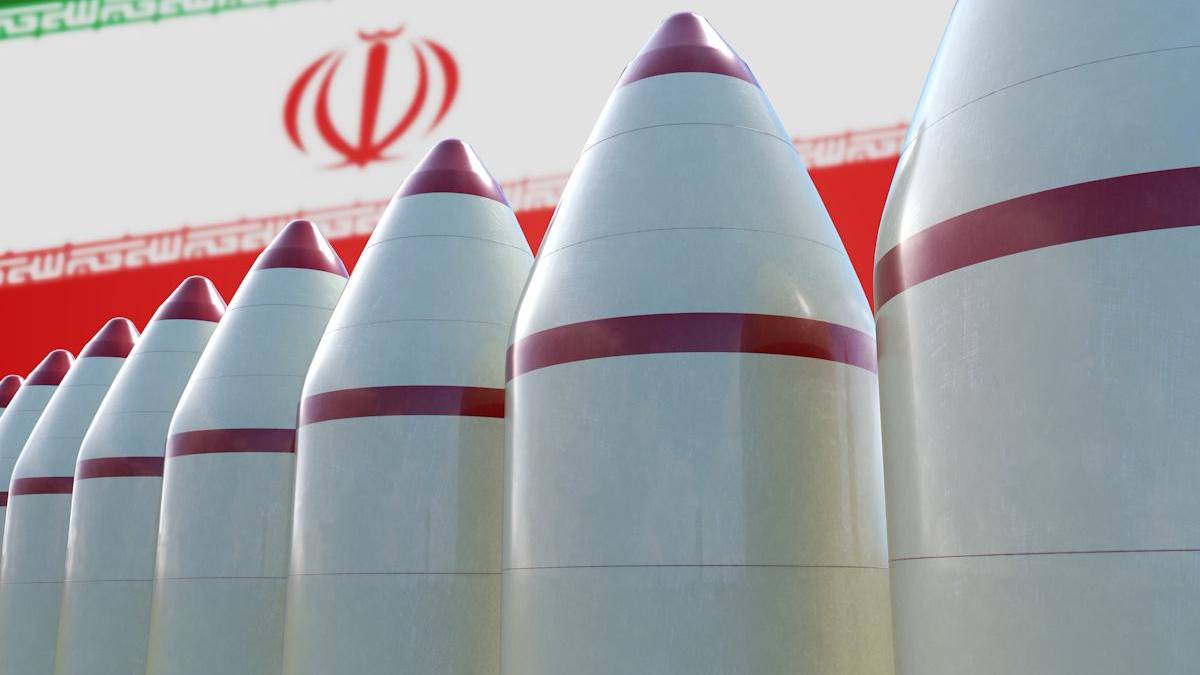 Row of white missiles with red tips and bands, lined up with Iran's flag and emblem in the background, blurred behind them, conveying a weapons display.