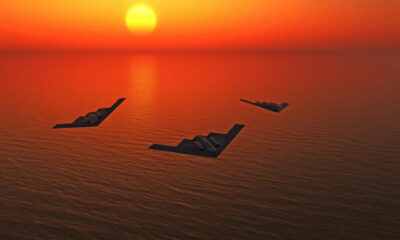 Three stealth bomber aircraft fly in a loose triangle over an orange sunset-lit ocean.