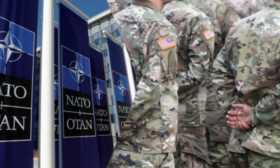 Soldiers in camouflage uniforms stand in line beside large NATO banners outside a building.