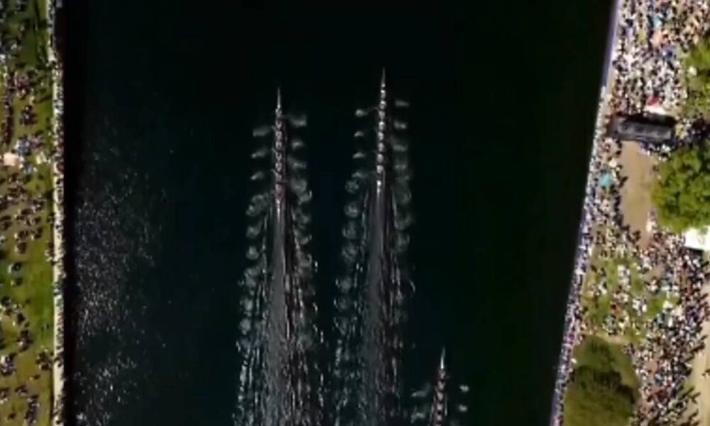 Aerial view of a rowing race on a dark river with multiple long boats and crowded shores on both sides.