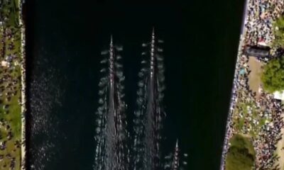 Aerial view of a rowing race on a dark river with multiple long boats and crowded shores on both sides.