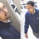 Split-screen surveillance image of a man with glasses in a dark jacket; left panel in an elevator, right panel walking in a hall with vending machines nearby