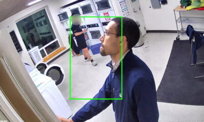 Frame from a security camera in a laundry room; foreground man in a blue jacket reaches toward a door, while another person near washers in the background is outlined by a green box.