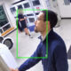 Frame from a security camera in a laundry room; foreground man in a blue jacket reaches toward a door, while another person near washers in the background is outlined by a green box.