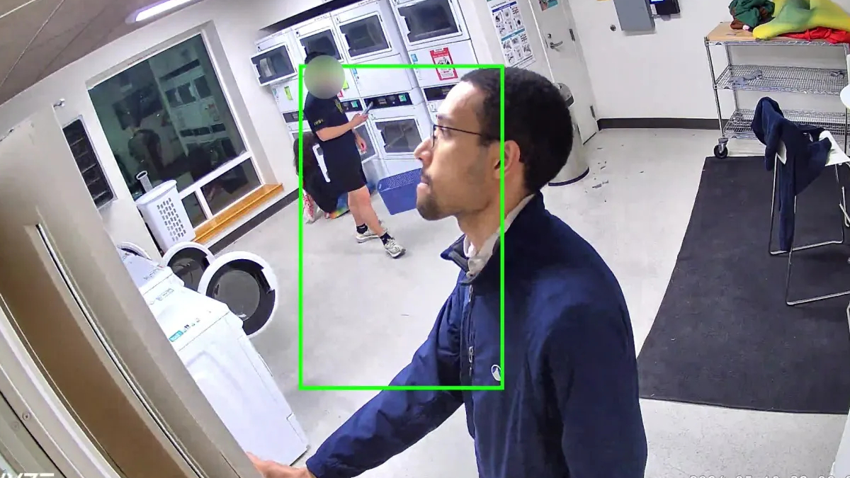 Frame from a security camera in a laundry room; foreground man in a blue jacket reaches toward a door, while another person near washers in the background is outlined by a green box.