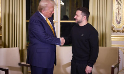 Two men shake hands in an opulent, gold-trimmed room with cream furniture and tall gold curtains in the background.