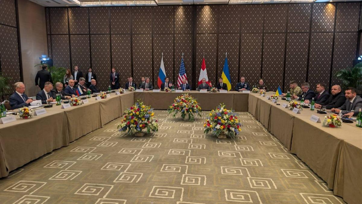 International diplomatic meeting with officials seated around a U-shaped beige table, flags of Russia, the US, Ukraine, Japan and others in the background, and floral centerpieces on the table edge.