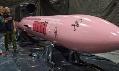 Pink rocket-shaped prop in a workshop, two men observing it, with black plastic sheeting and equipment surrounding the scene.
