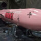 Pink rocket-shaped prop in a workshop, two men observing it, with black plastic sheeting and equipment surrounding the scene.