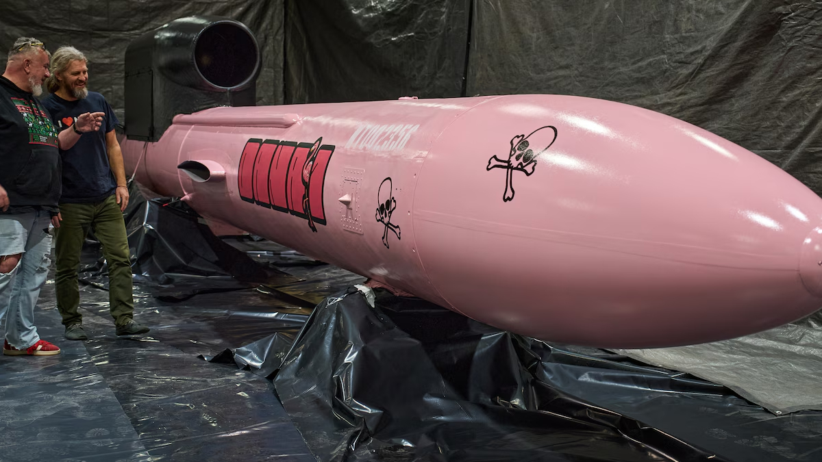 Pink rocket-shaped prop in a workshop, two men observing it, with black plastic sheeting and equipment surrounding the scene.