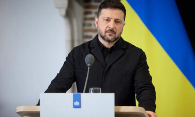 Man in a black jacket speaks at a podium with a microphone, with a Ukrainian flag backdrop behind him.