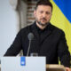 Man in a black jacket speaks at a podium with a microphone, with a Ukrainian flag backdrop behind him.