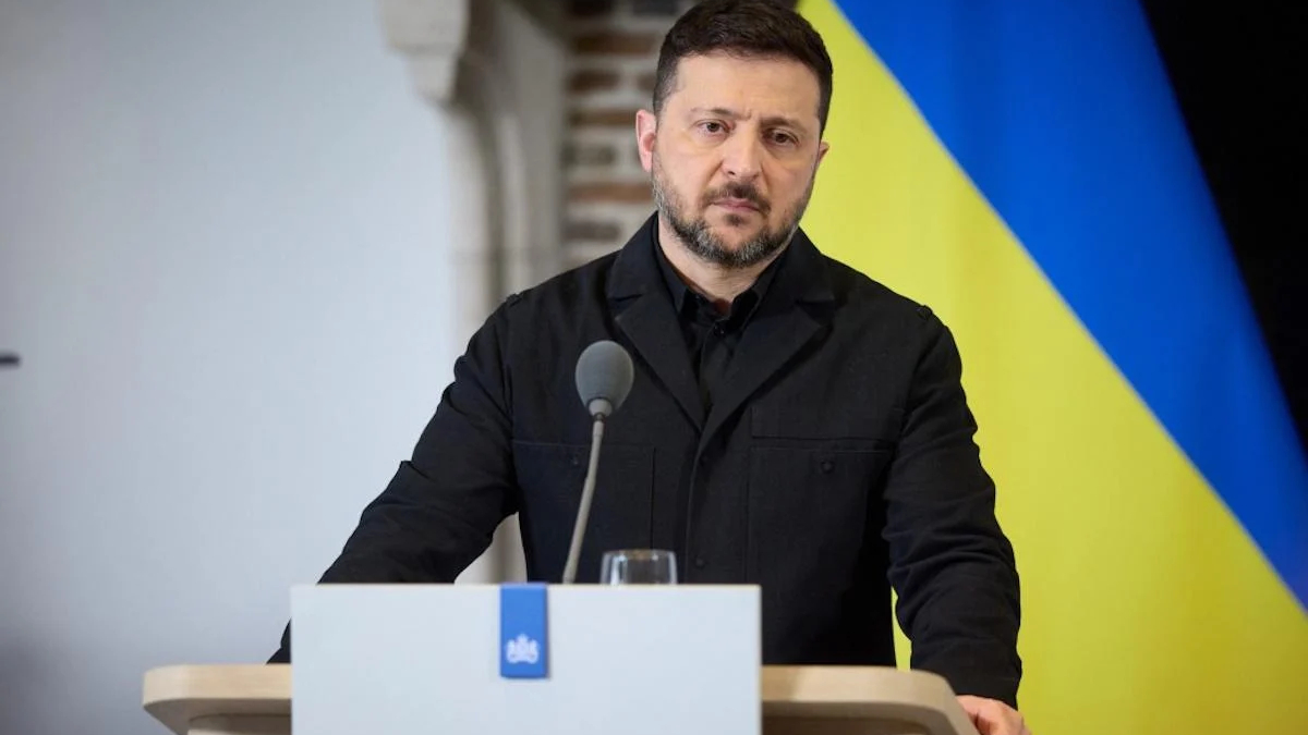 Man in a black jacket speaks at a podium with a microphone, with a Ukrainian flag backdrop behind him.