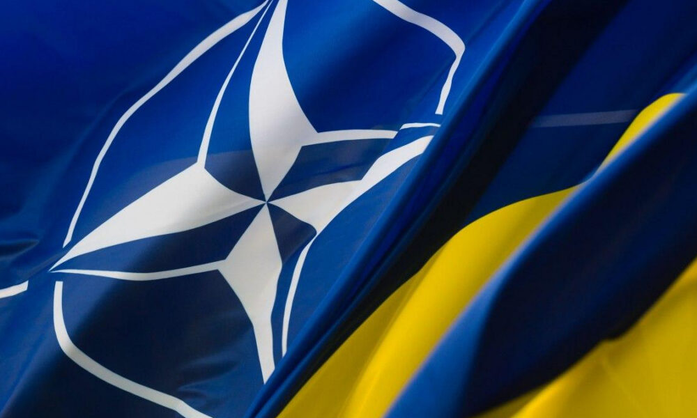 Flags of NATO and Ukraine waving together: blue flag with white NATO emblem beside a blue-and-yellow Ukrainian flag, symbolizing alliance and support.