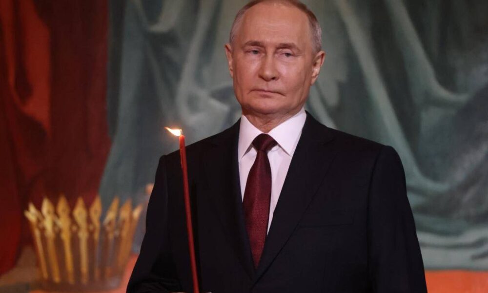 Man in a dark suit and red tie holding a lit red candle at a ceremony, with a ceremonial backdrop.