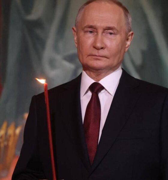 Man in a dark suit and red tie holding a lit red candle at a ceremony, with a ceremonial backdrop.