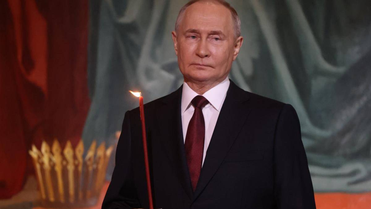 Man in a dark suit and red tie holding a lit red candle at a ceremony, with a ceremonial backdrop.