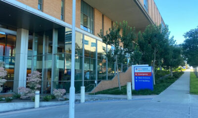 Exterior of a modern medical building with glass walls, trees, and a blue sign directing to emergency and main entrance. (Providence)