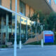 Exterior of a modern medical building with glass walls, trees, and a blue sign directing to emergency and main entrance. (Providence)