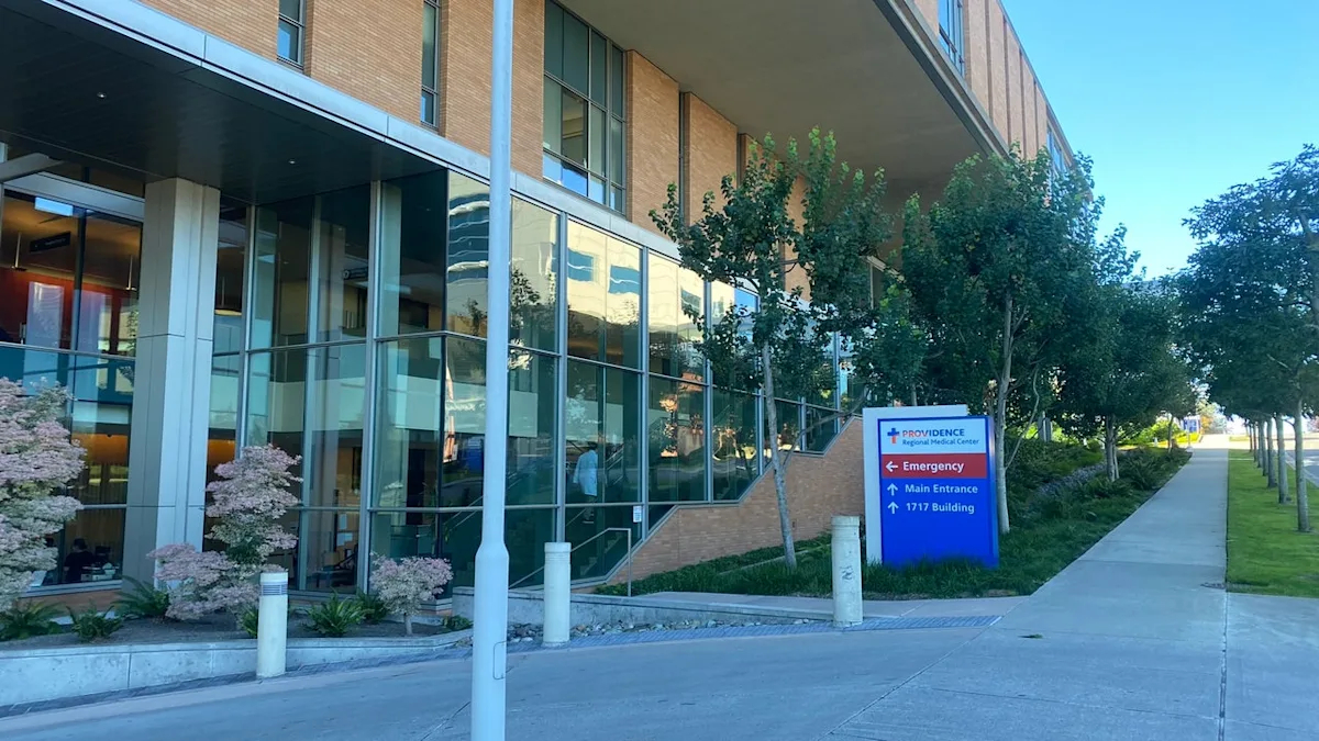 Exterior of a modern medical building with glass walls, trees, and a blue sign directing to emergency and main entrance. (Providence)