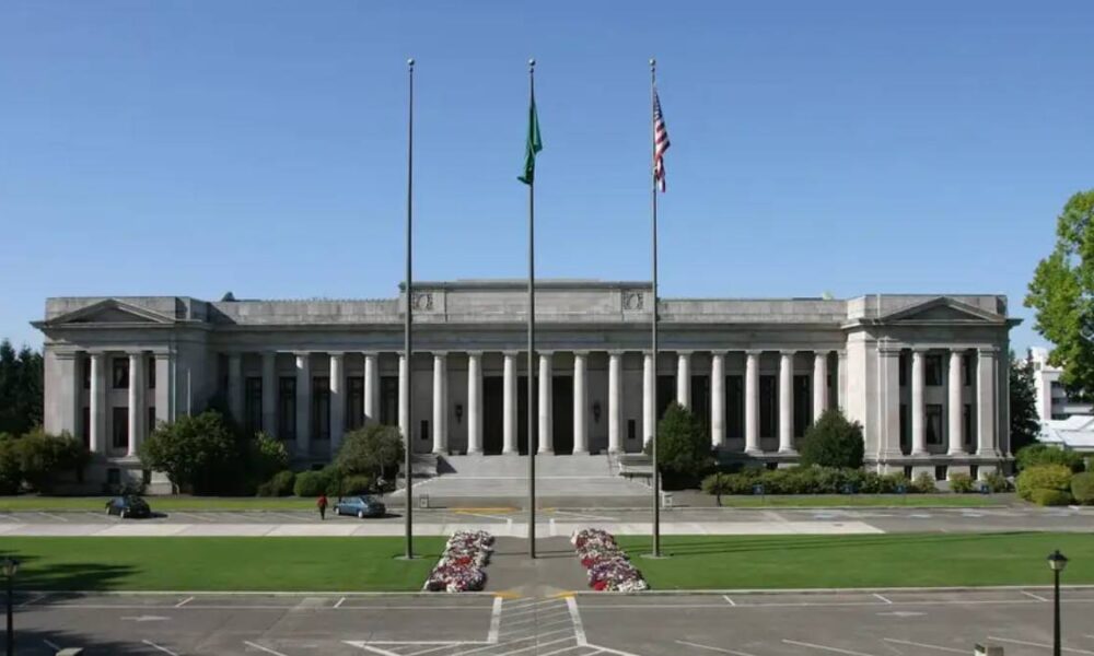 Neoclassical government building with tall columns and three flagpoles in front, green lawn and flower beds.