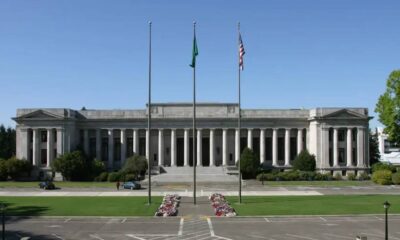 Neoclassical government building with tall columns and three flagpoles in front, green lawn and flower beds.