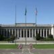 Neoclassical government building with tall columns and three flagpoles in front, green lawn and flower beds.