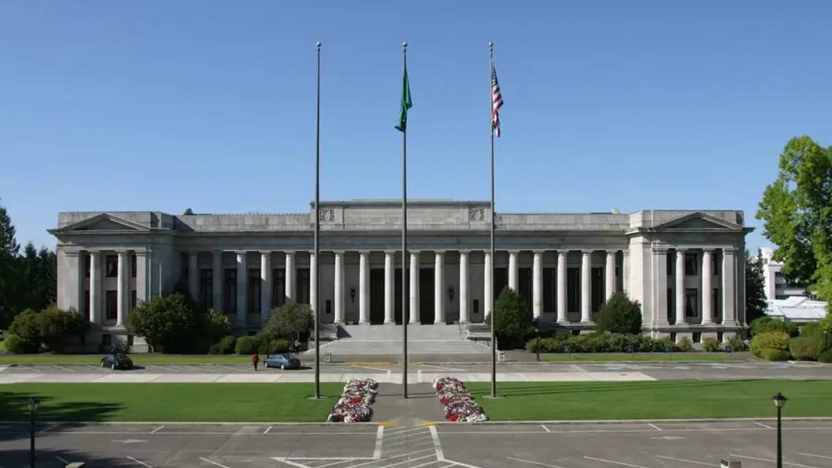 Neoclassical government building with tall columns and three flagpoles in front, green lawn and flower beds.