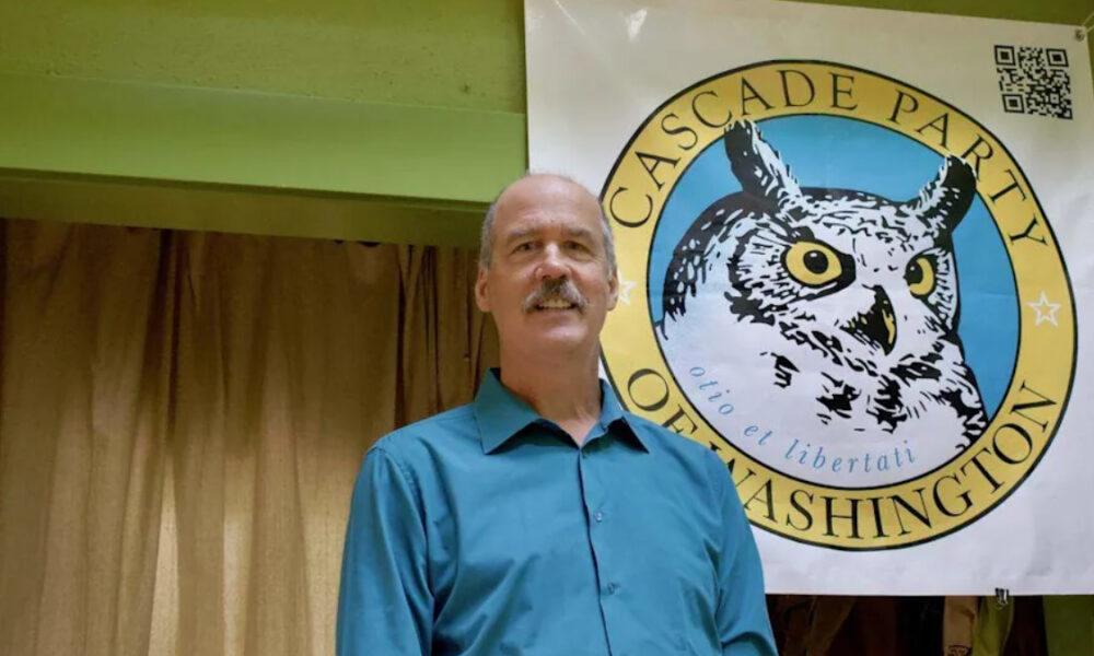Man in a teal shirt standing in front of a Cascade Party banner with a large owl emblem behind him