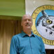 Man in a teal shirt standing in front of a Cascade Party banner with a large owl emblem behind him