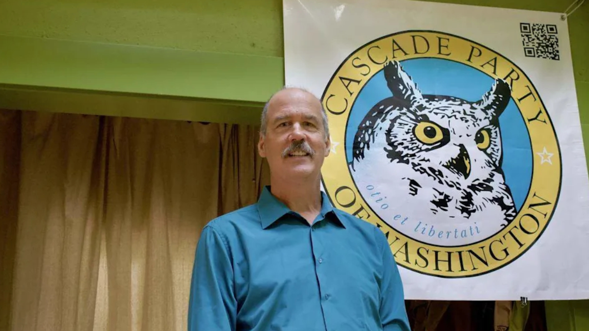 Man in a teal shirt standing in front of a Cascade Party banner with a large owl emblem behind him