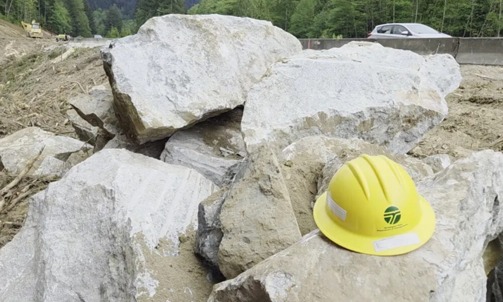 Yellow construction hard hat resting on large rocks at a worksite with a road and forest in the background.