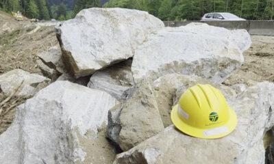 Yellow construction hard hat resting on large rocks at a worksite with a road and forest in the background.