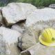 Yellow construction hard hat resting on large rocks at a worksite with a road and forest in the background.