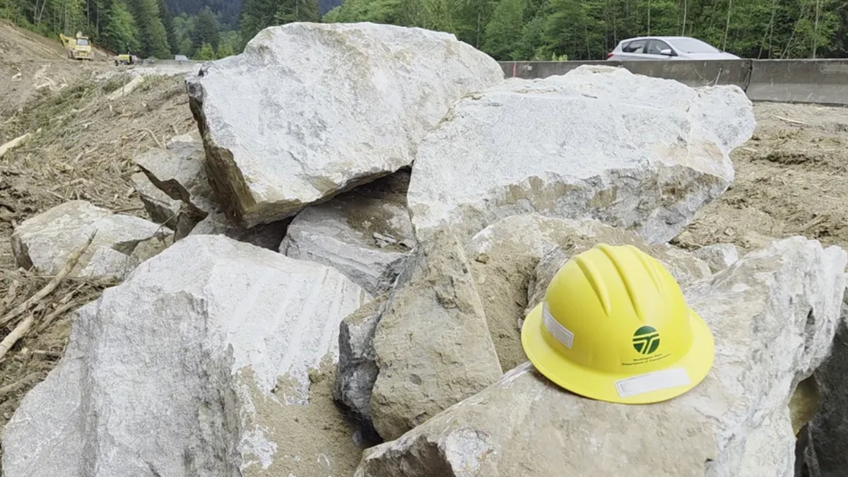 Yellow construction hard hat resting on large rocks at a worksite with a road and forest in the background.