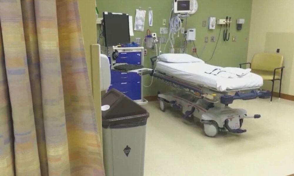 Hospital patient room with a wheeled stretcher, medical devices on the wall, and a beige chair to the right.