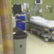 Hospital patient room with a wheeled stretcher, medical devices on the wall, and a beige chair to the right.