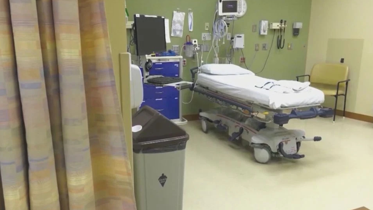 Hospital patient room with a wheeled stretcher, medical devices on the wall, and a beige chair to the right.
