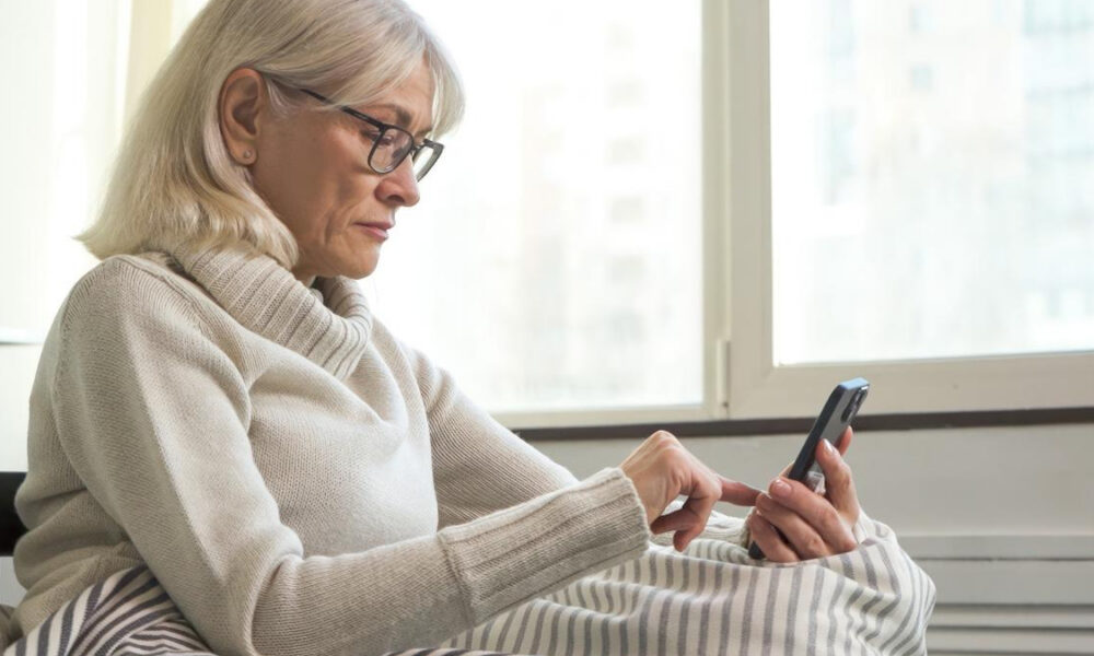 Older woman with glasses in a cozy sweater using a smartphone in bed by a window.