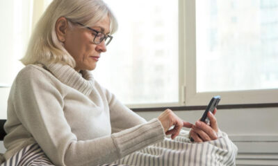 Older woman with glasses in a cozy sweater using a smartphone in bed by a window.