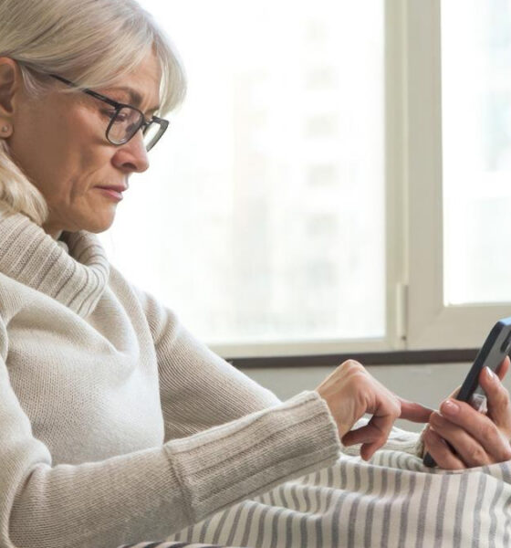 Older woman with glasses in a cozy sweater using a smartphone in bed by a window.