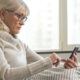 Older woman with glasses in a cozy sweater using a smartphone in bed by a window.