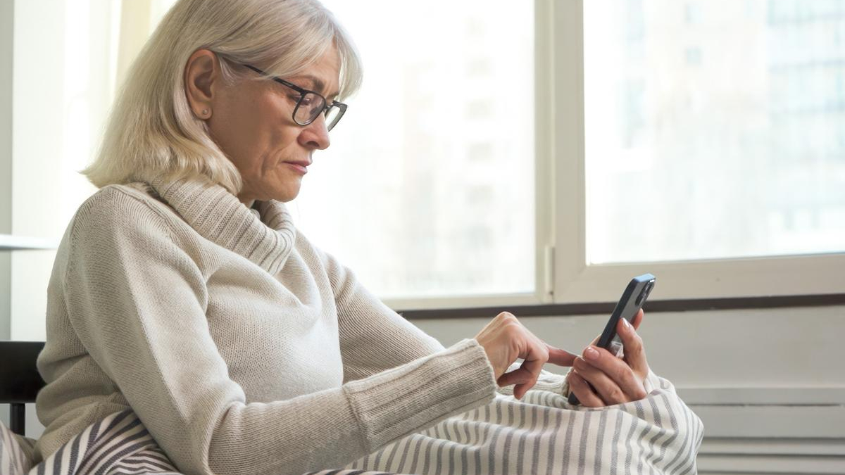 Older woman with glasses in a cozy sweater using a smartphone in bed by a window.