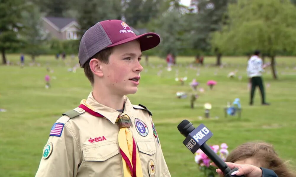 Boy scout in uniform being interviewed by a reporter in a grassy park filled with small flower decorations in the background.