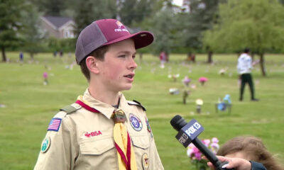 Boy scout in uniform being interviewed by a reporter in a grassy park filled with small flower decorations in the background.